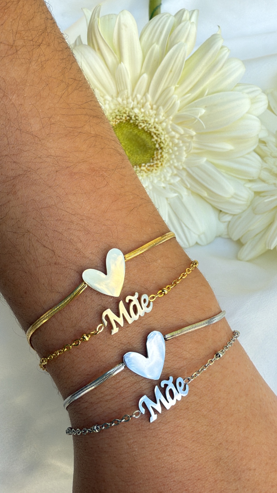 Dois conjuntos de pulseiras dourada e prateada com corações e palavra 'Mãe' e flor branca ao fundo