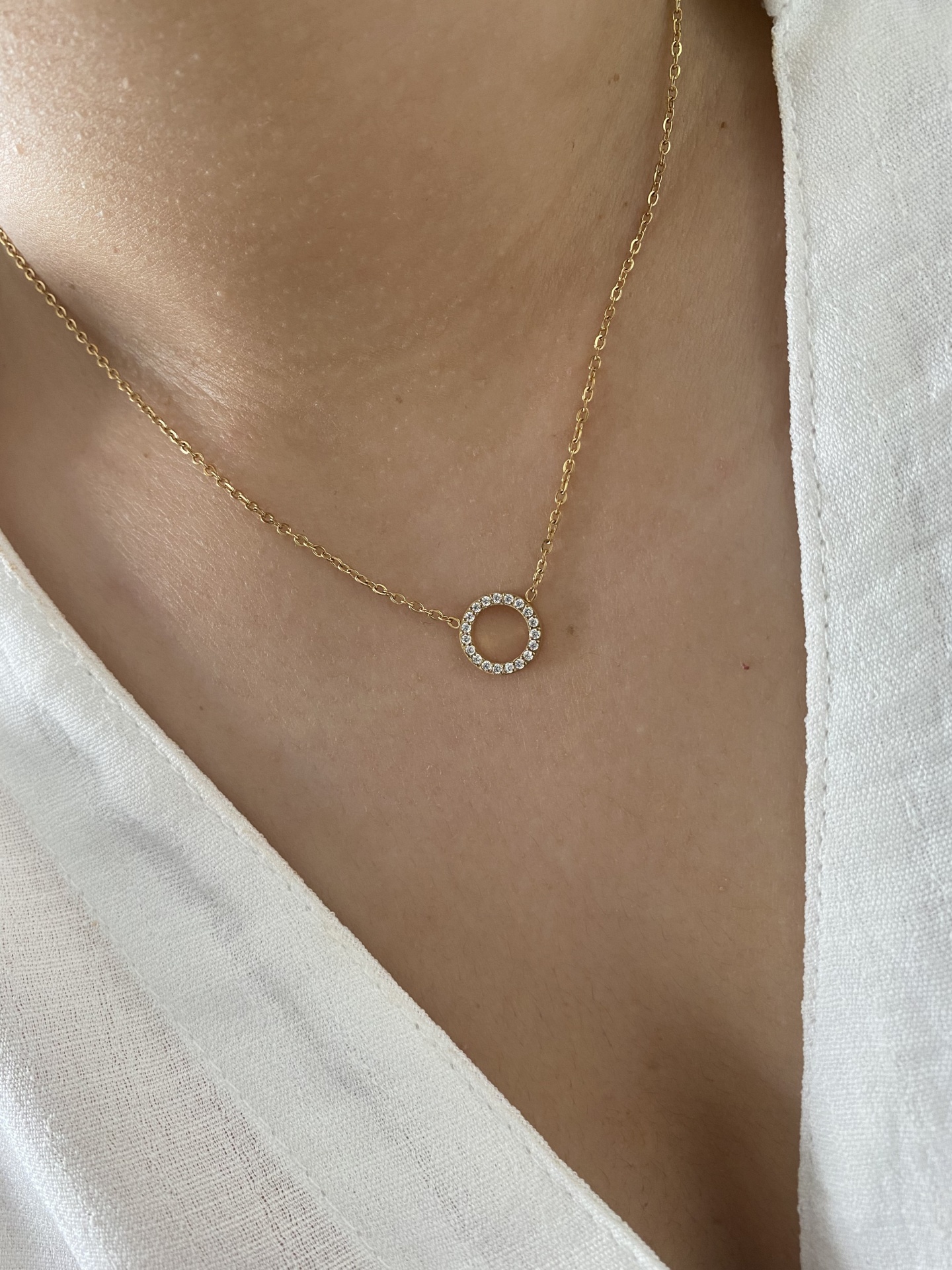 Colar dourado com pendente circular de cristais num pescoço com camisa branca