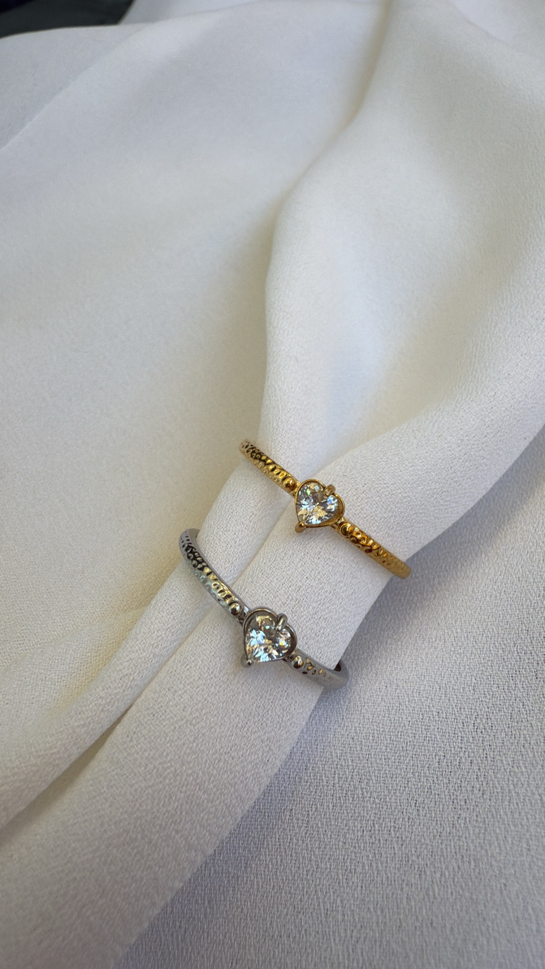 Dois anéis delicados com pedras em forma de coração, um dourado e outro prateado, sobre tecido branco.