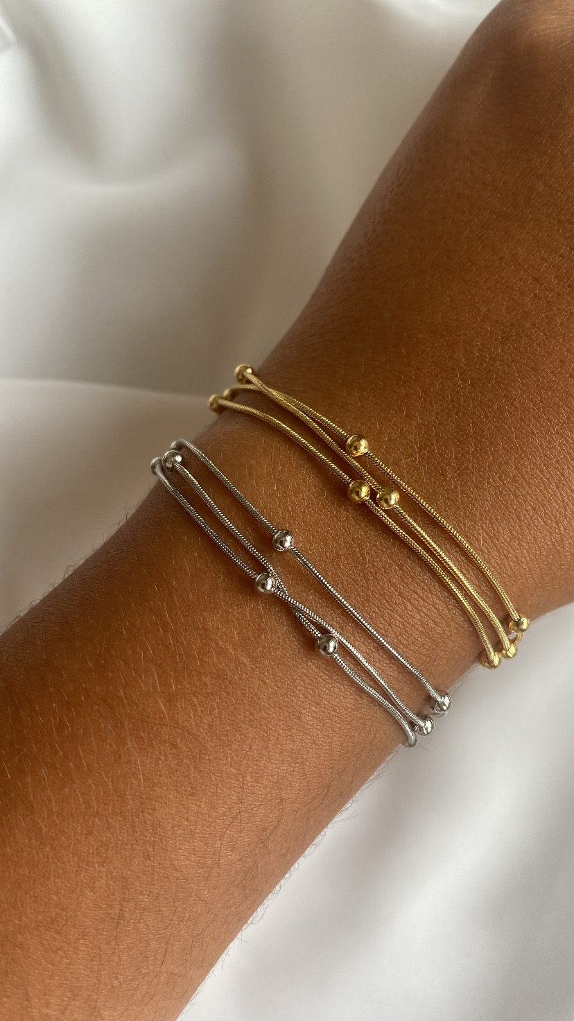 Pulseiras finas douradas e prateadas com bolas metálicas no pulso.