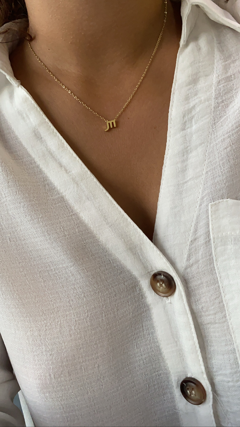 Colar dourado com letra m em corrente fina, camisa branca com botões castanhos.