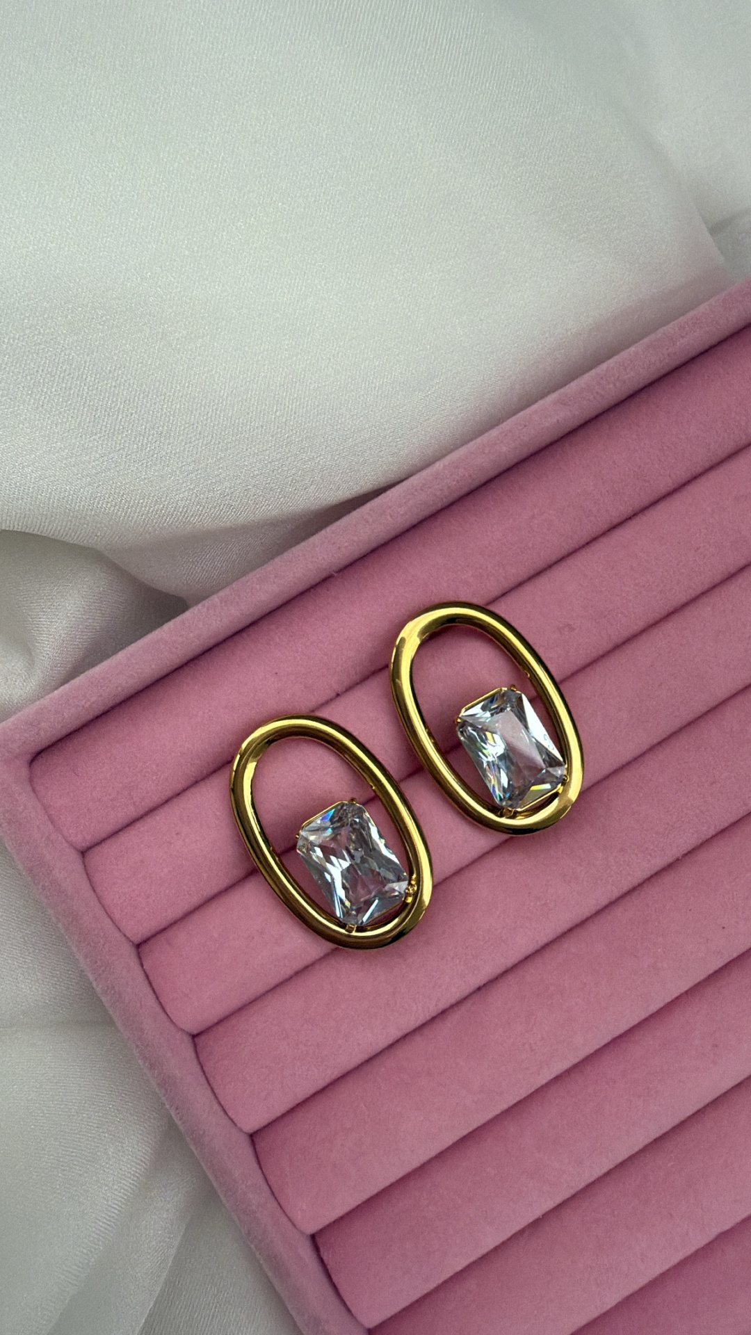 Brincos dourados ovais com pedras transparentes em caixa rosa.
