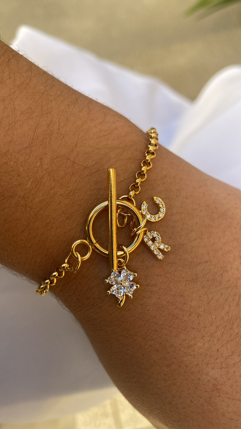 Pulseira dourada com letras C e R em pedras brilhantes num pulso.