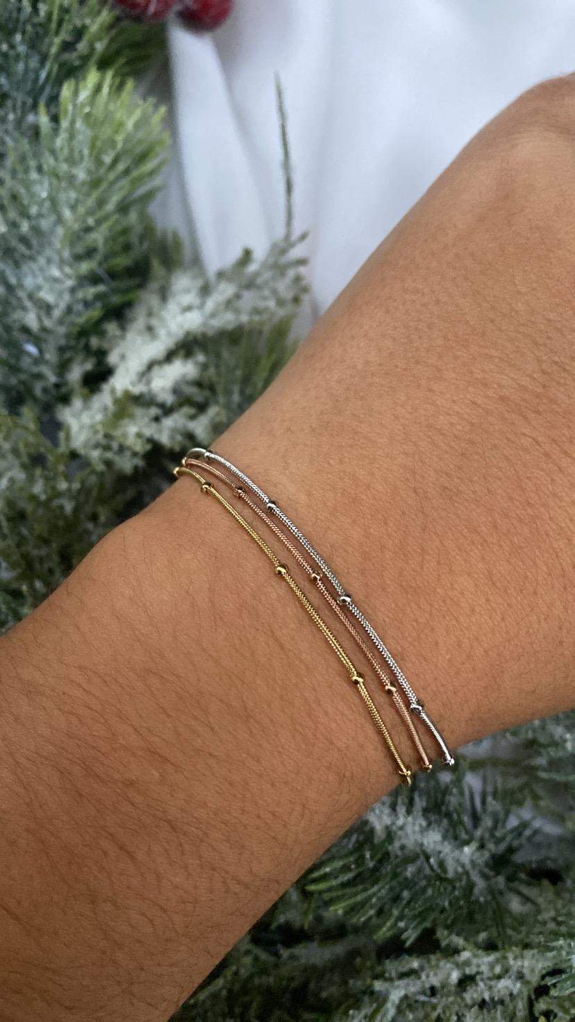 Pulseira fina dourada e prateada com pequenas esferas no pulso junto a folhagem verde festiva