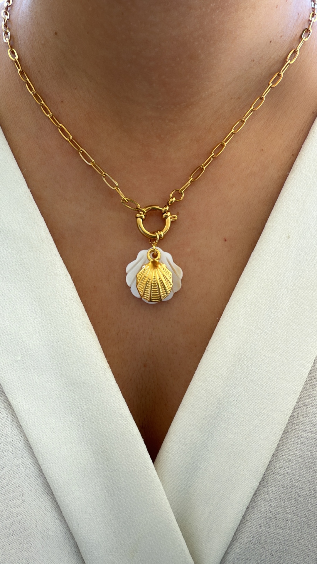 Colar dourado com pendente de concha e fundo branco, usado ao redor do pescoço.