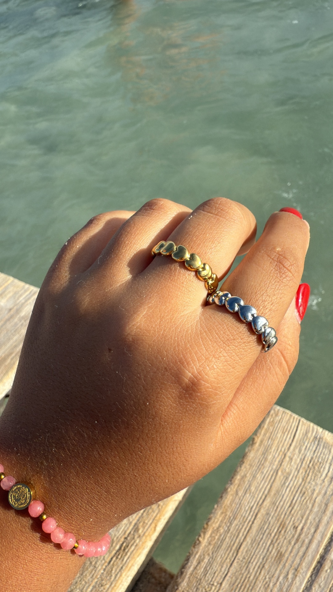Mão com dois anéis de coração dourado e prateado e pulseira de contas rosas junto à água