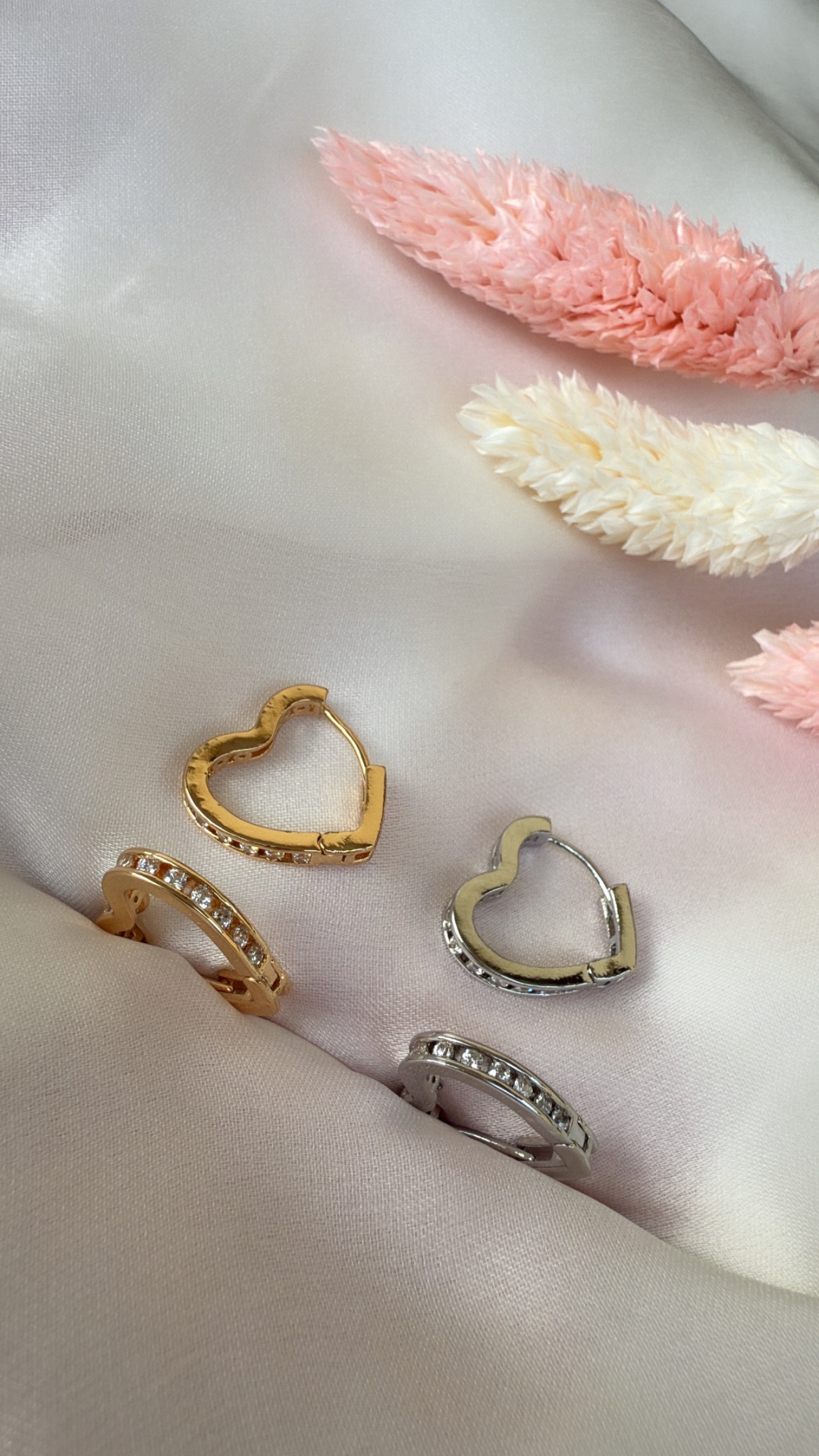 Brincos dourados e prateados de coração e com pedras sobre tecido acetinado creme com flores secas rosa e creme