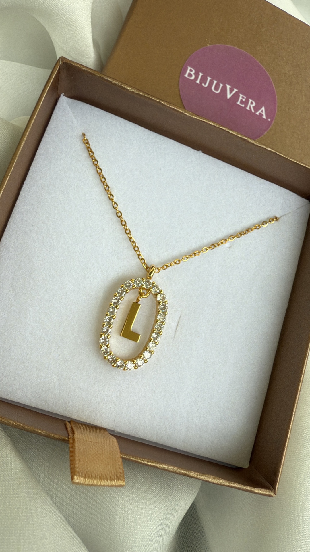 Colar dourado com pendente oval de pedras brilhantes e letra L numa caixa castanha com interior branco e etiqueta BIJUVERA
