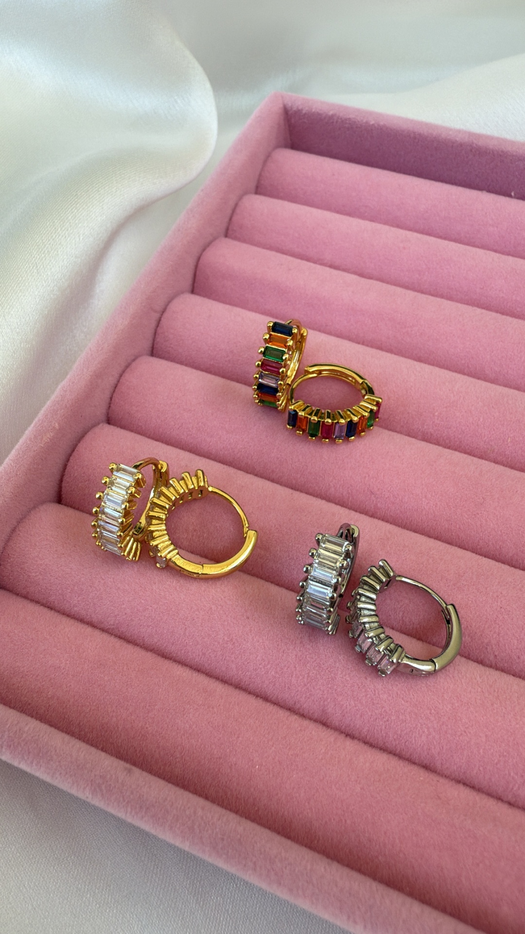 Três pares de brincos em aro com pedras coloridas e transparentes numa caixa rosa acolchoada.