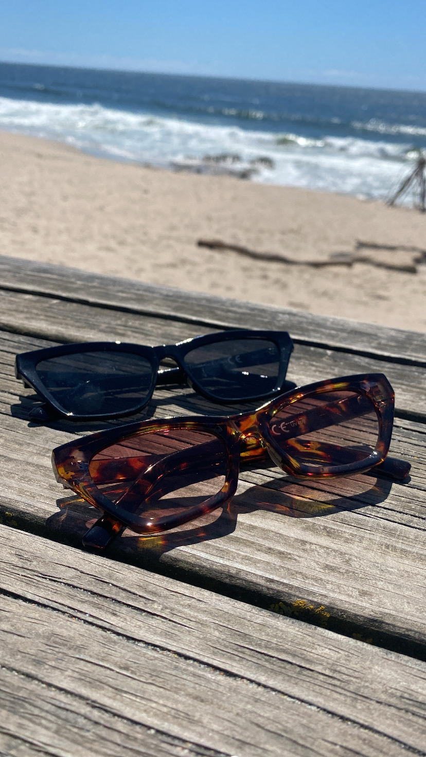 Dois pares de óculos de sol numa mesa de madeira com vista para a praia e mar.