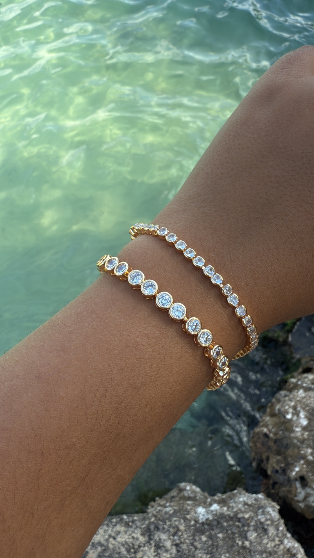 Pulseiras douradas com pedras transparentes num pulso junto a água e pedras