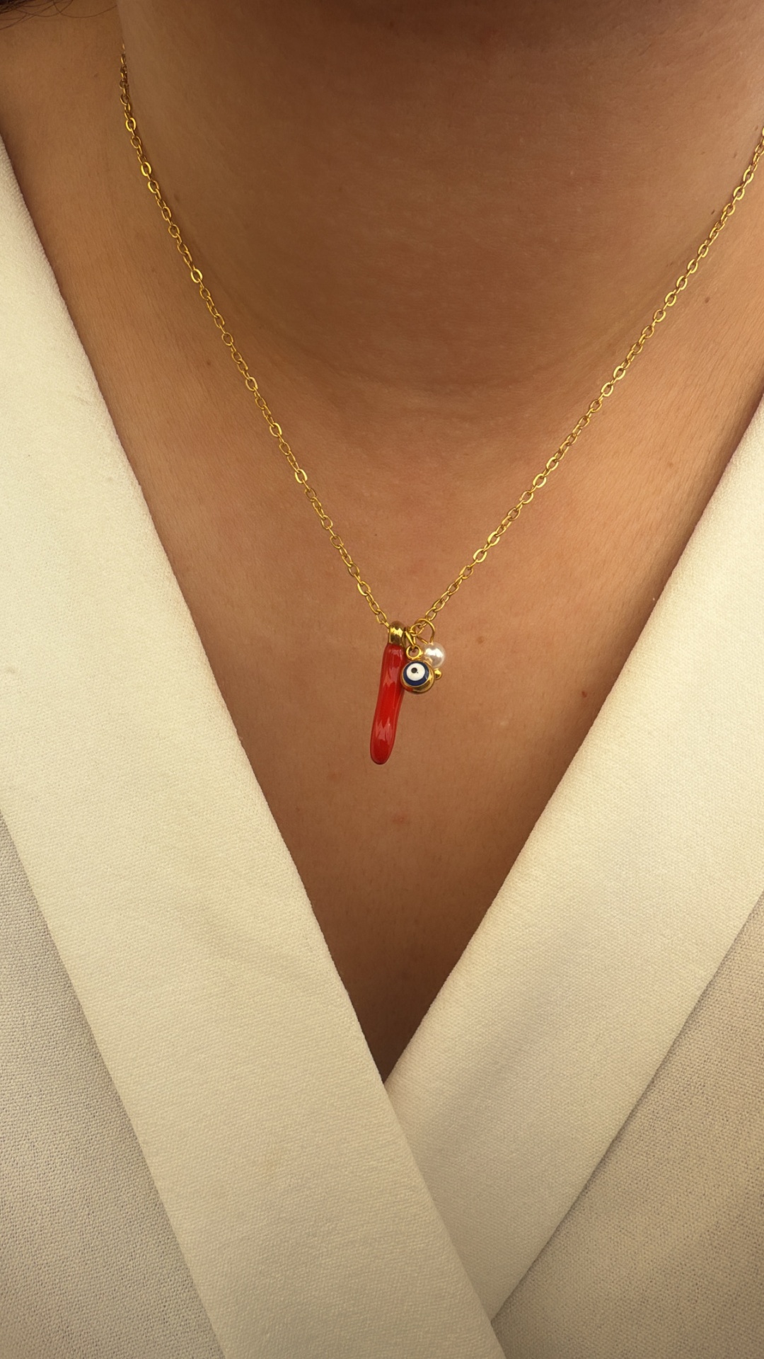 Colar dourado com pendentes vermelho, olho azul e pérola sobre pele e roupa branca.