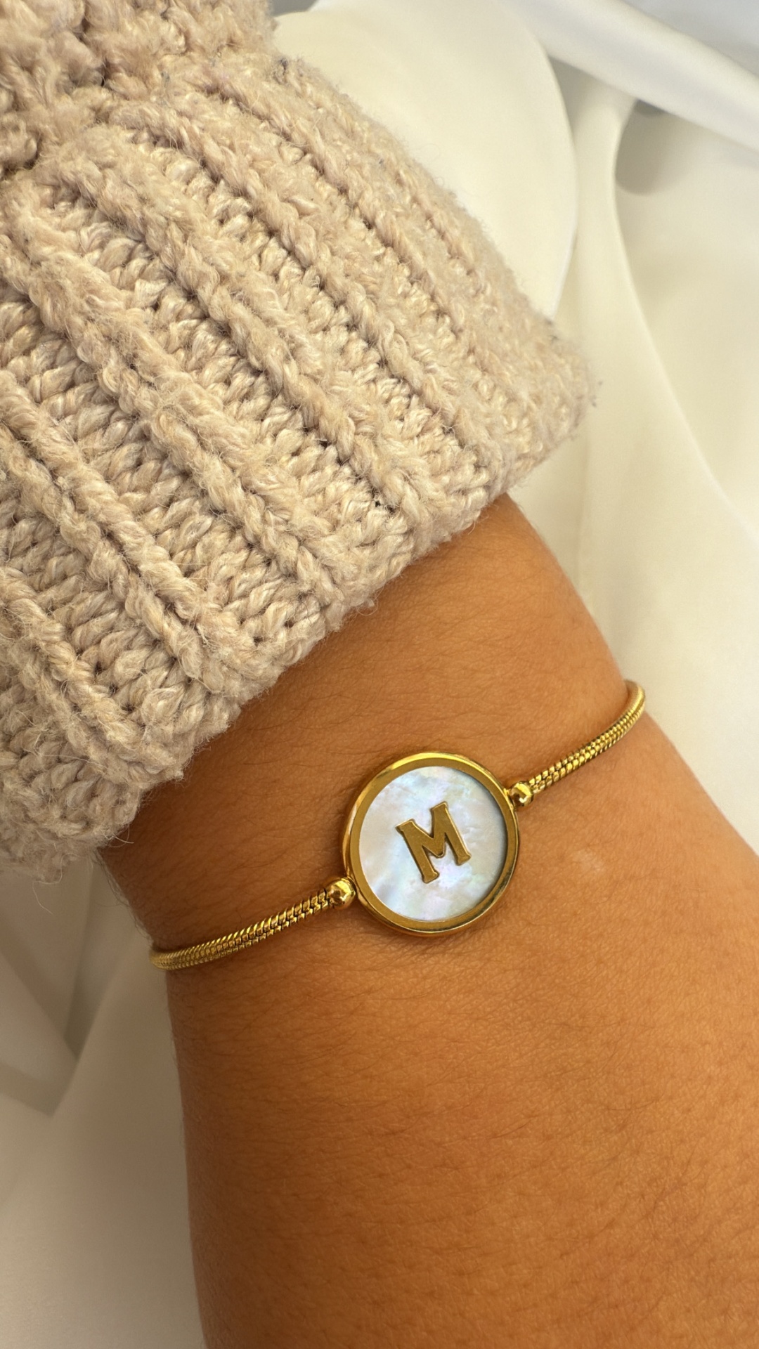 Pulseira dourada com medalha redonda e letra M ao centro num pulso com manga de malha bege.
