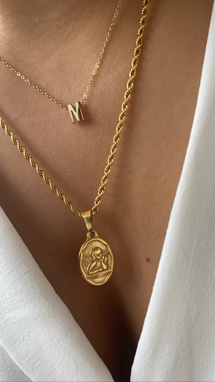 Colares dourados com pendente oval e letra M sobre pele e camisa branca