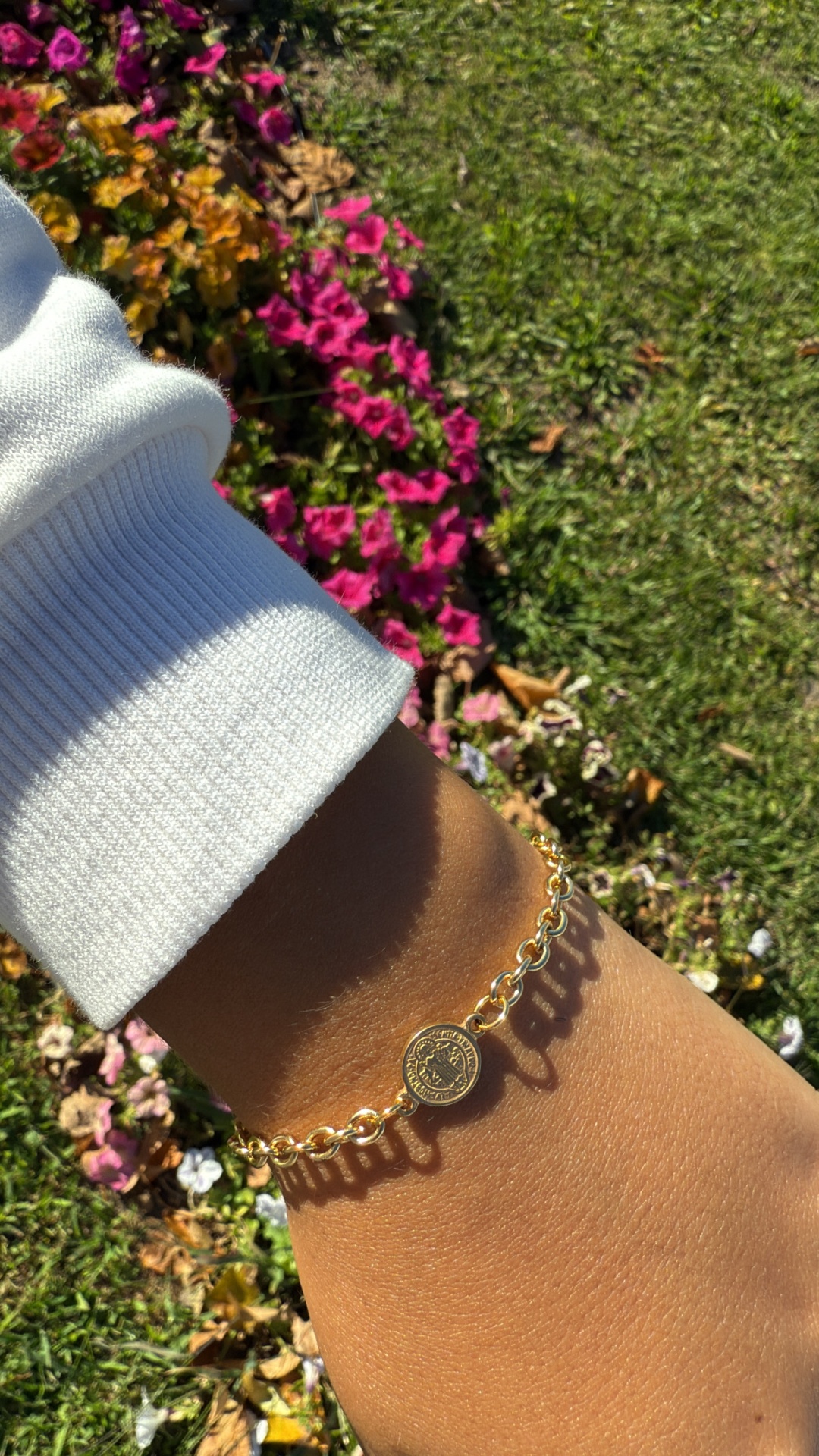 Pulseira dourada com pendente circular em pulso contra flores e relvado