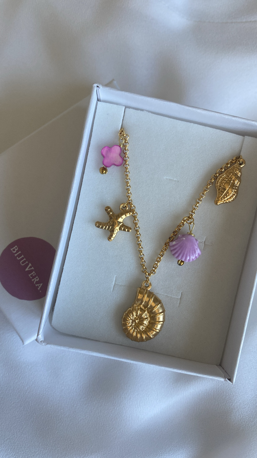 Colar dourado com pendentes de concha, estrela do mar e detalhes rosa numa caixa branca com etiqueta 'BIJURERA'.