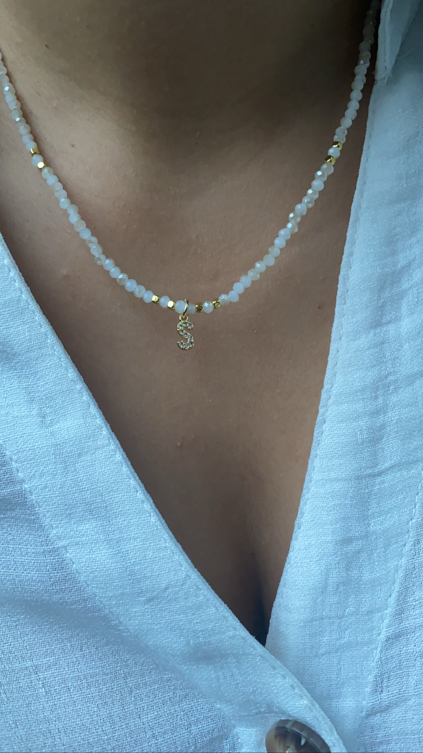 Colar com contas brancas e douradas e pendente em forma de S, usado com camisa branca.