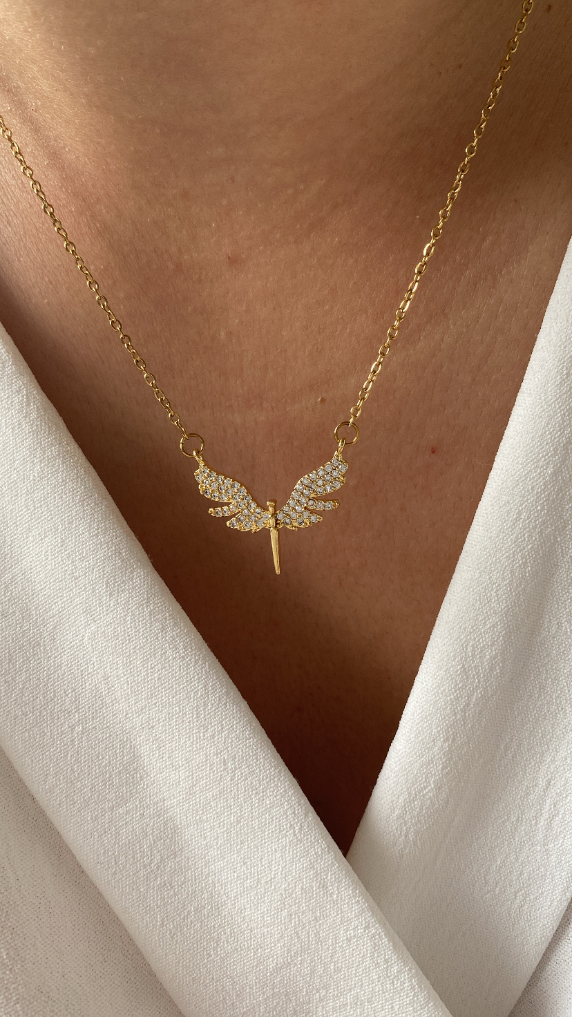 Colar dourado com pendente de asas brilhantes em pele clara e vestido branco.