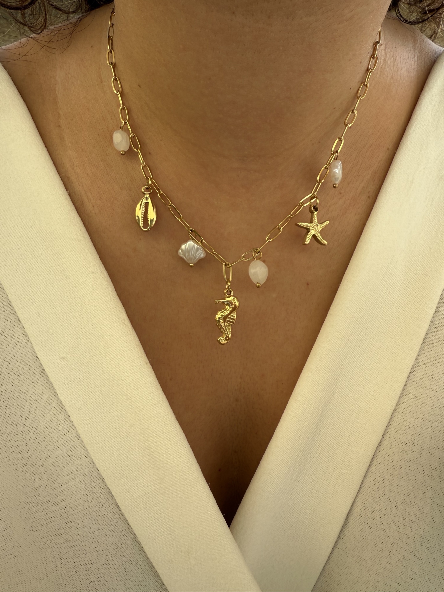 Colar dourado com pendentes marinhos e pedras brancas em pescoço de pessoa com blusa creme