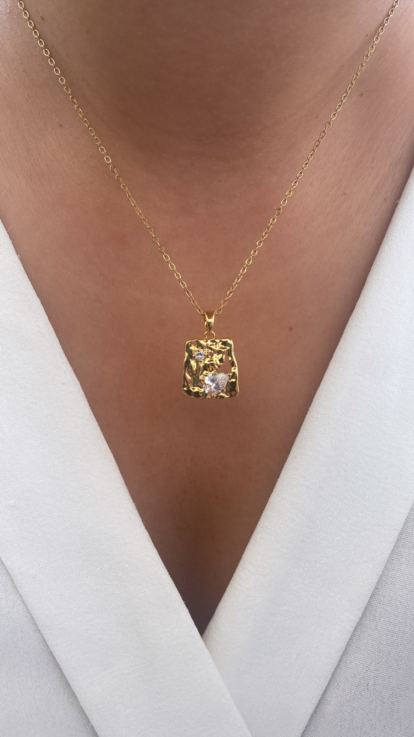 Colar dourado com pendente quadrado e pedra transparente sobre camisa branca