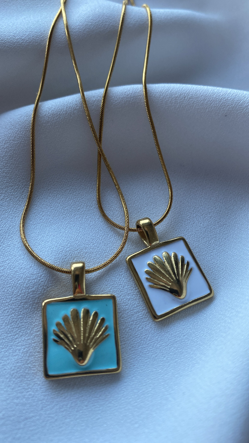 Dois colares dourados com pendentes quadrados azul claro e branco com conchas douradas em relevo sobre tecido branco