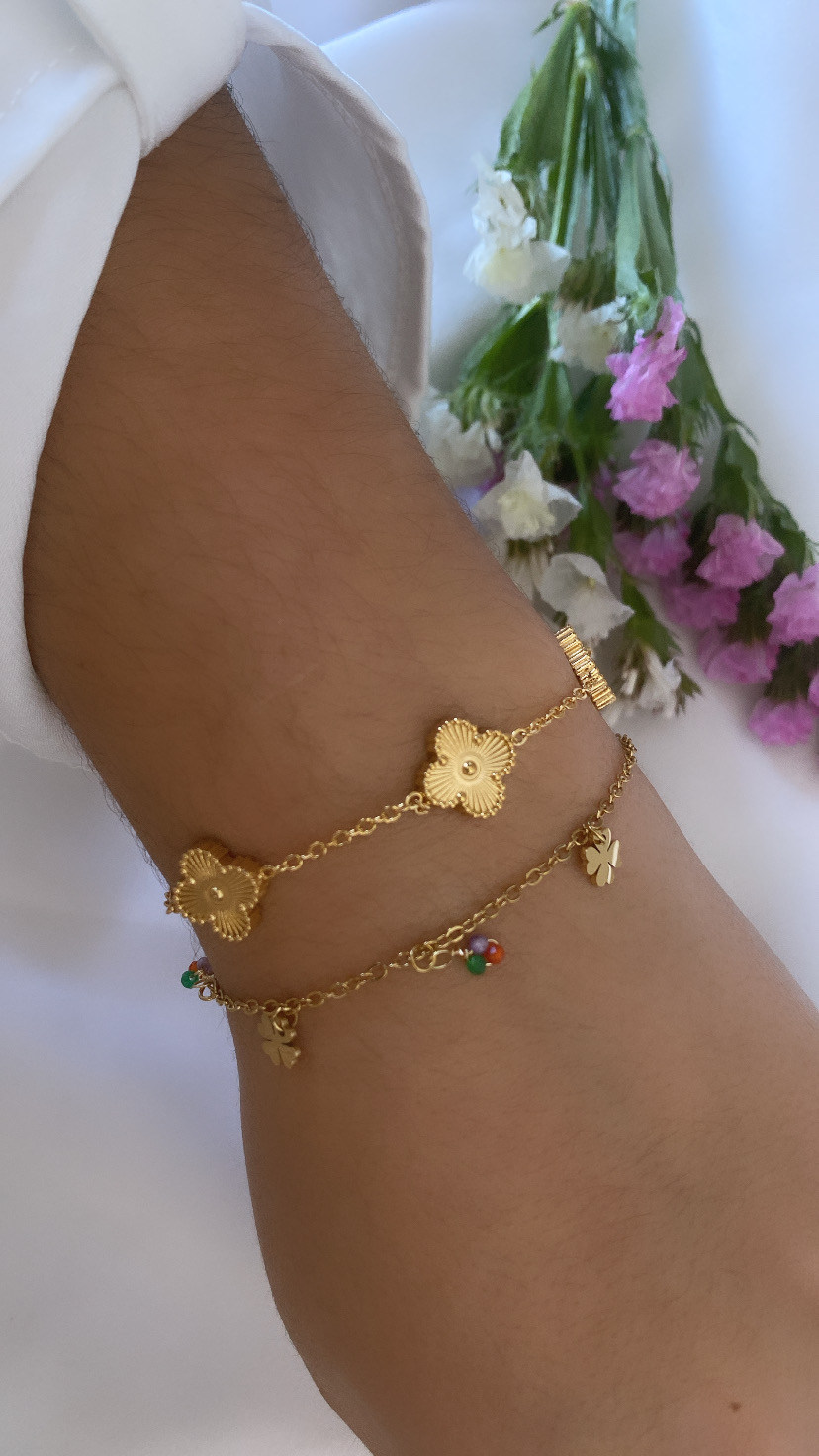 Pulseira dourada com flores e pendentes coloridos num pulso com camisa branca e flores ao fundo