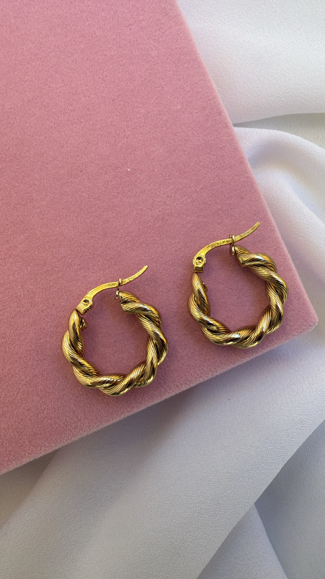Brincos de argola torcidos metálicos dourados sobre fundo rosa aveludado e branco.