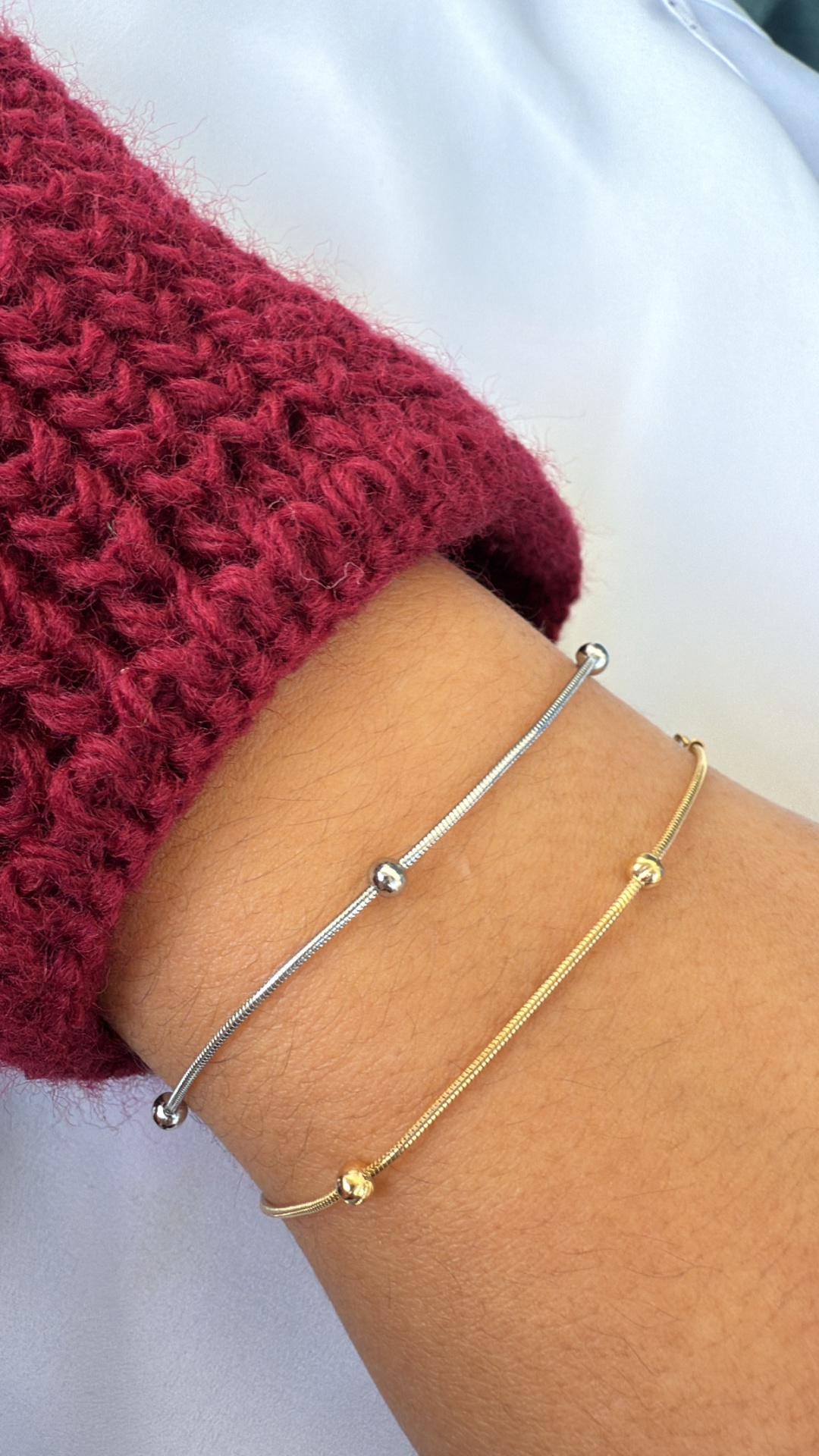 Pulseiras finas em prata e ouro com contas redondas num pulso. Roupas bordô e branco ao fundo.