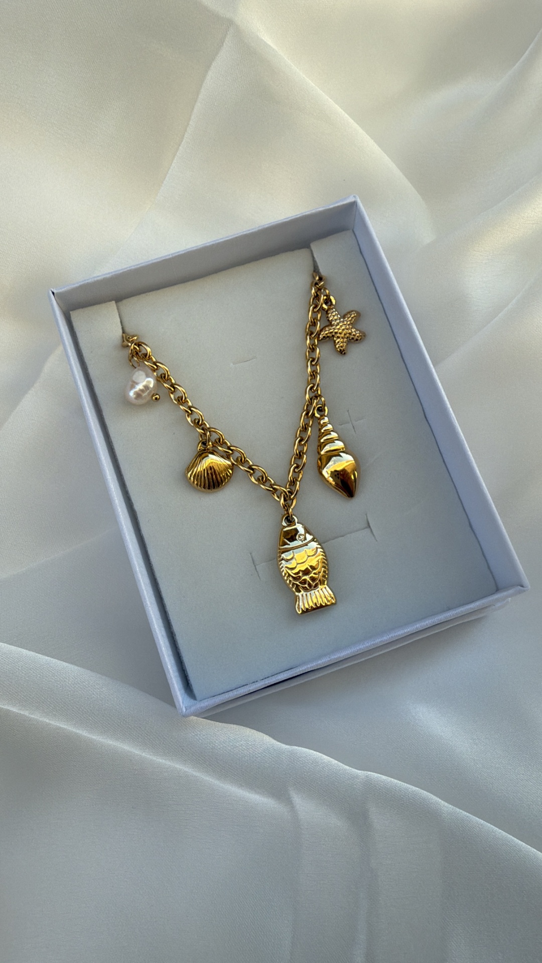 Colar dourado com pendentes marinhos numa caixa cinzenta sobre tecido branco