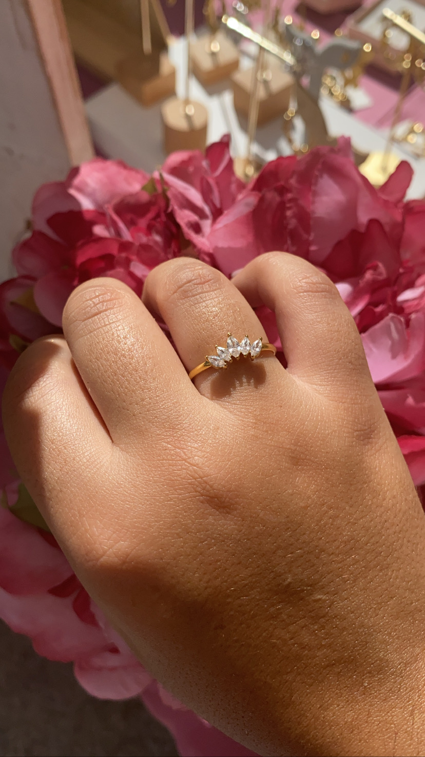 Anel dourado com cristais em forma de folhas num dedo com flores cor-de-rosa ao fundo