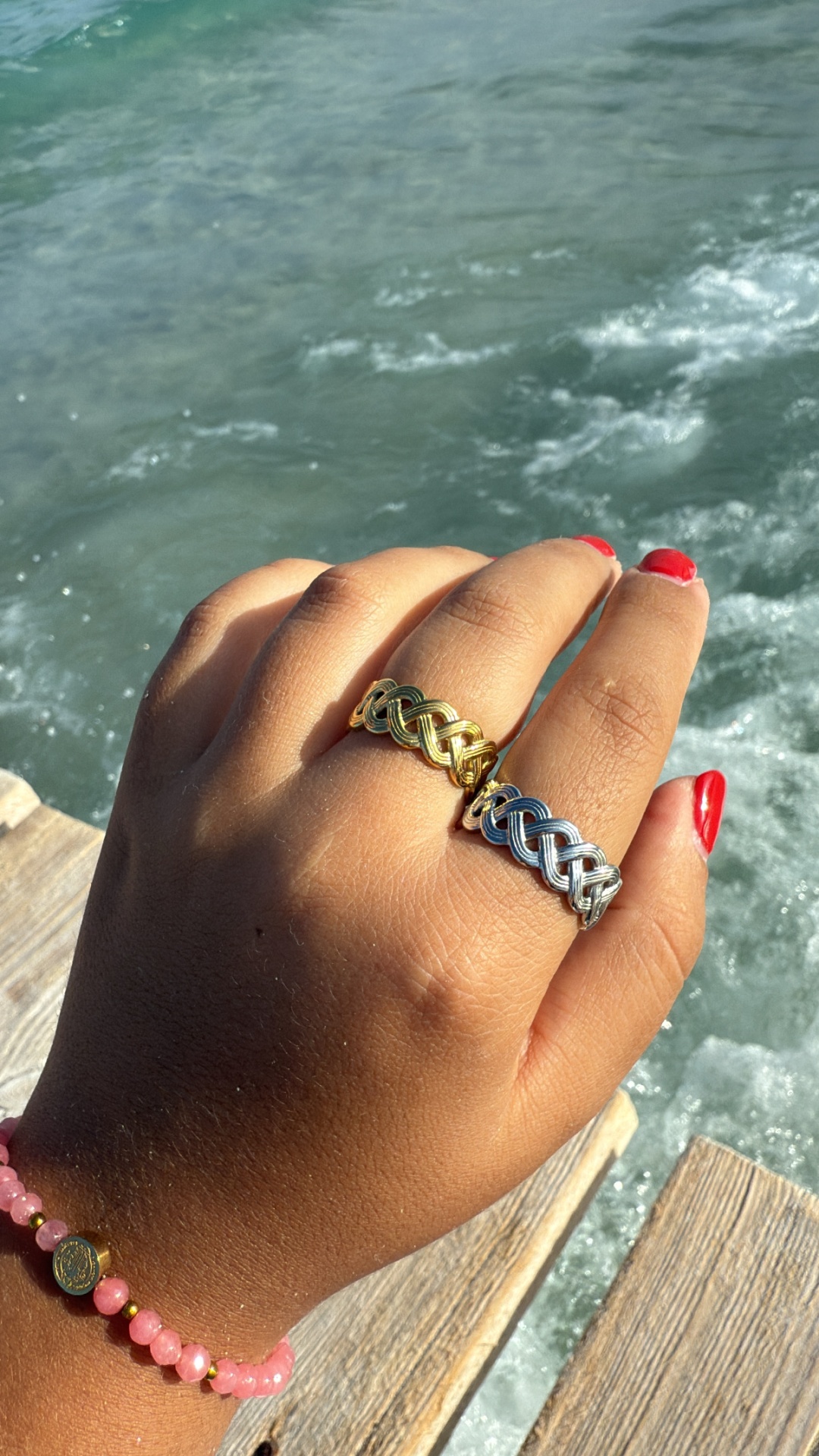 Mão com anéis dourado e prateado e pulseira rosa junto à água
