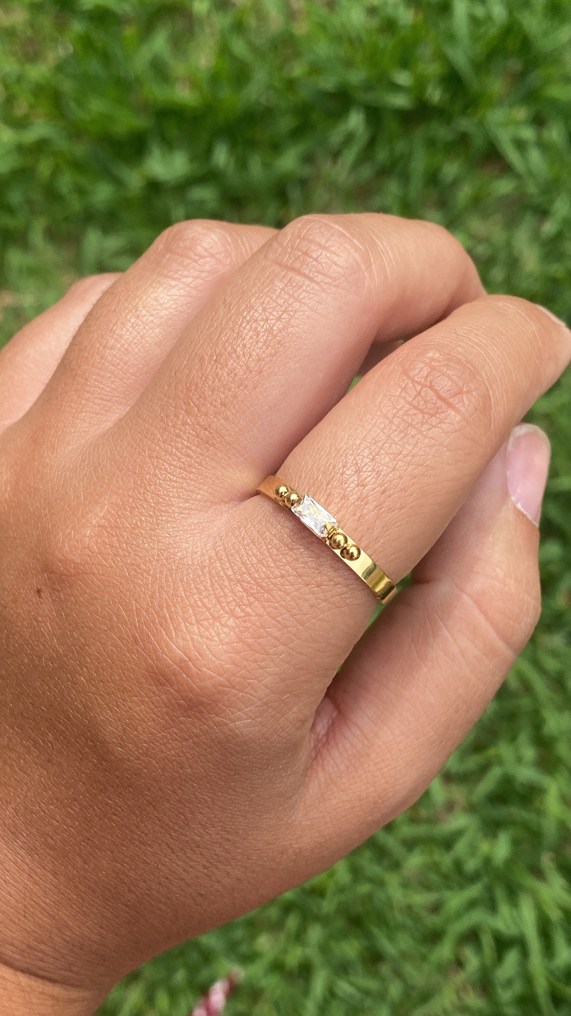 Anel dourado com pedra transparente e detalhes redondos numa mão sobre fundo de relva verde