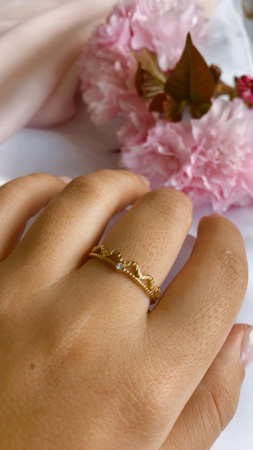 Anel dourado com design de coroa e pedra transparente num dedo com flores cor de rosa ao fundo