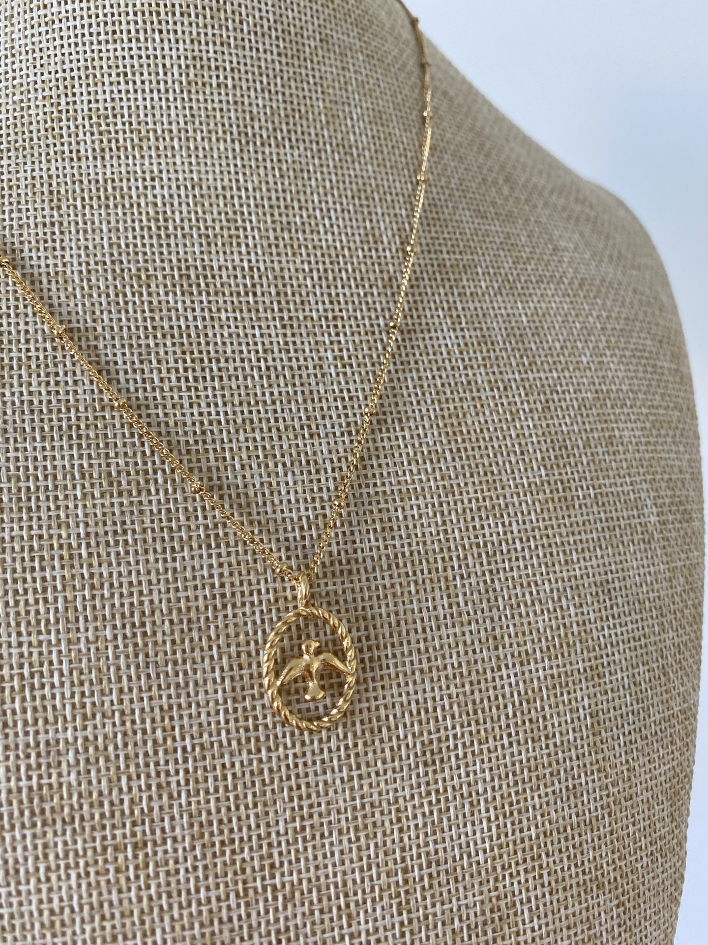 Colar dourado com pendente circular trançado e folhas numa base de tecido bege