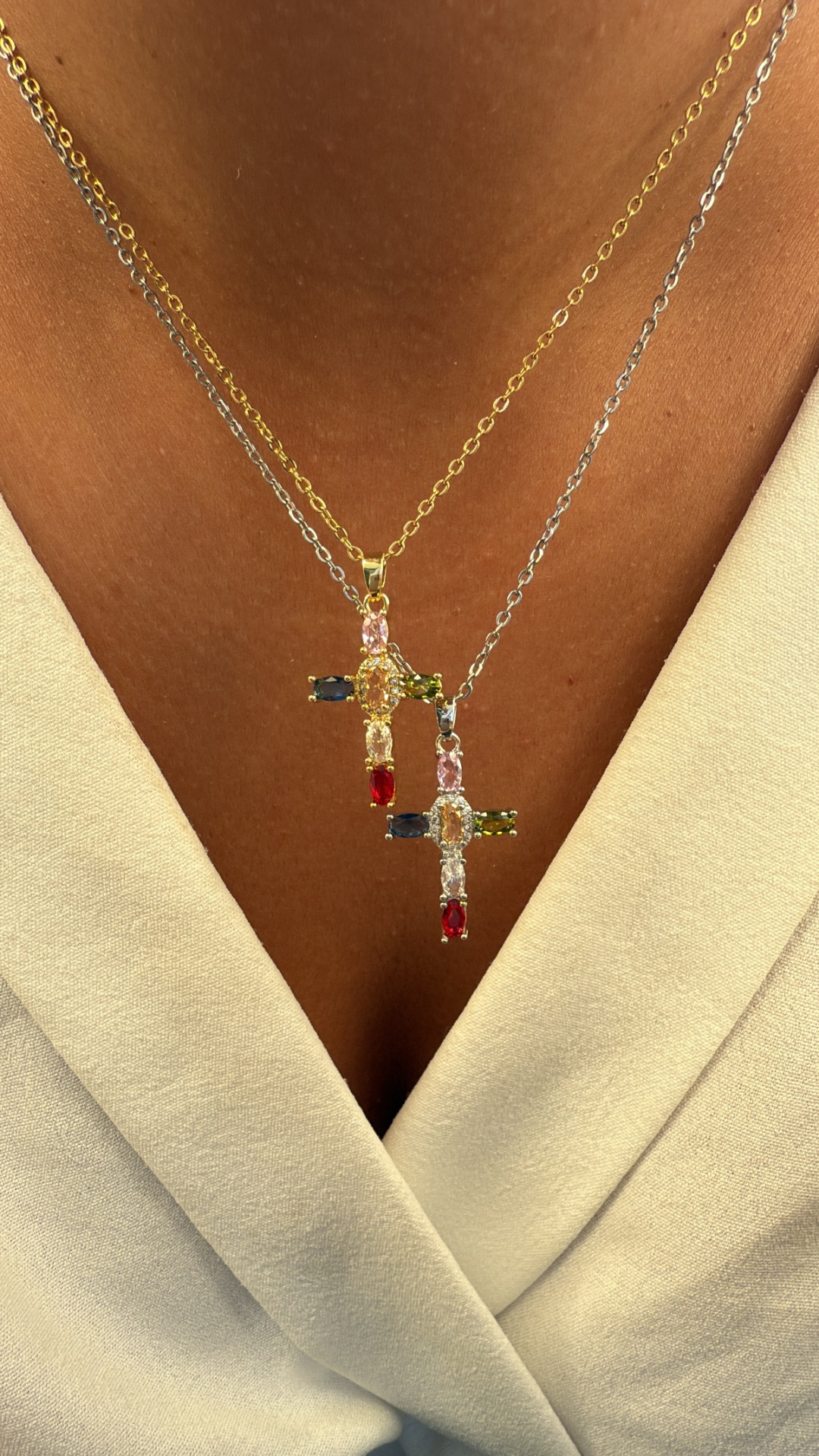 Dois colares com pendentes cruz gemas coloridas, correntes dourada e prateada em pele vestindo roupa branca.