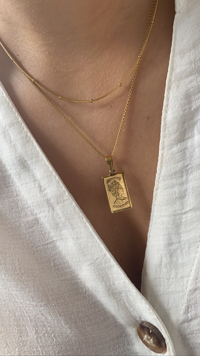 Colar duplo dourado com pendente retangular gravado, sobre blusa branca de linho.