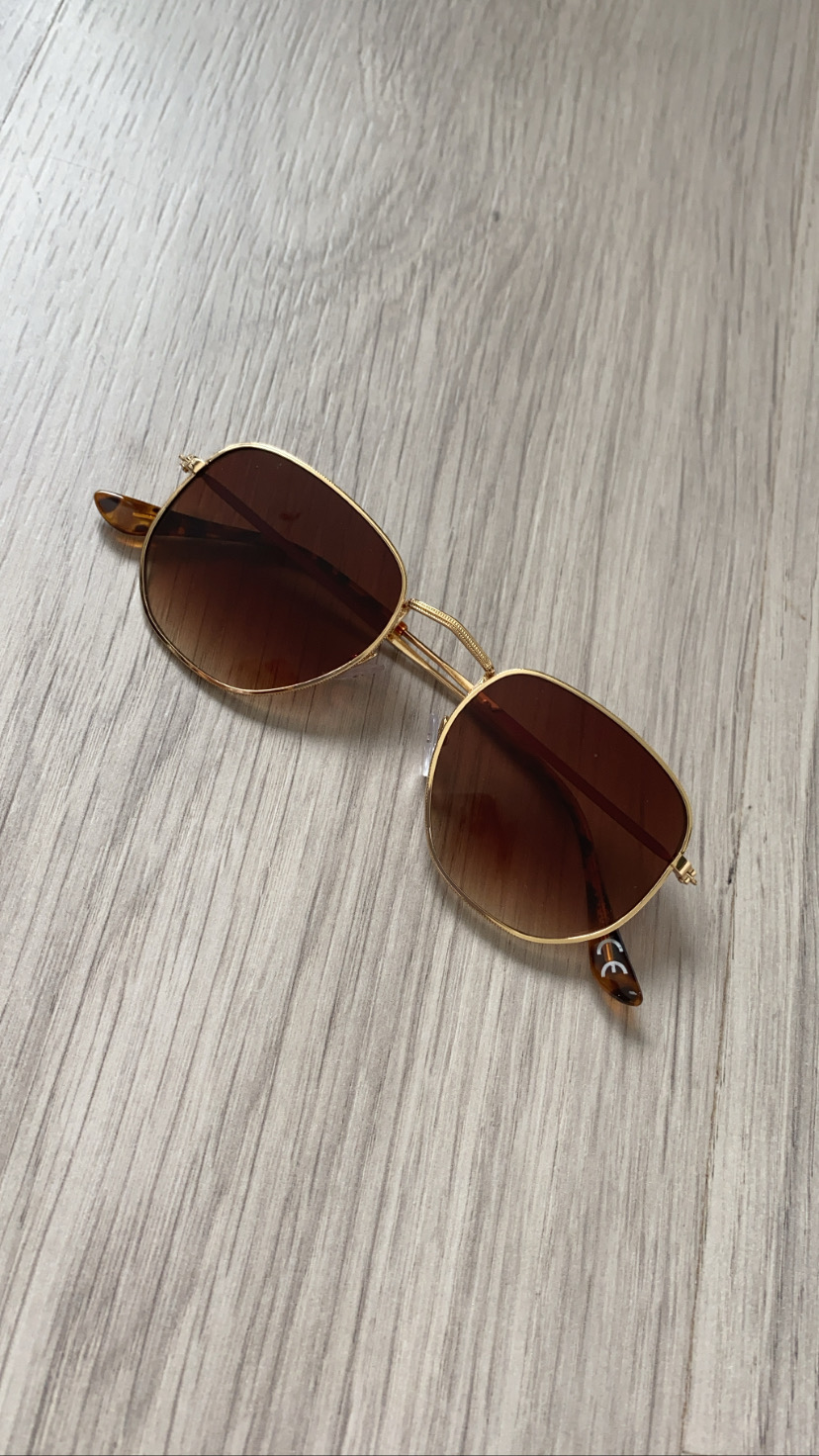 Óculos de sol com armação dourada e lentes castanho degradê sobre madeira clara