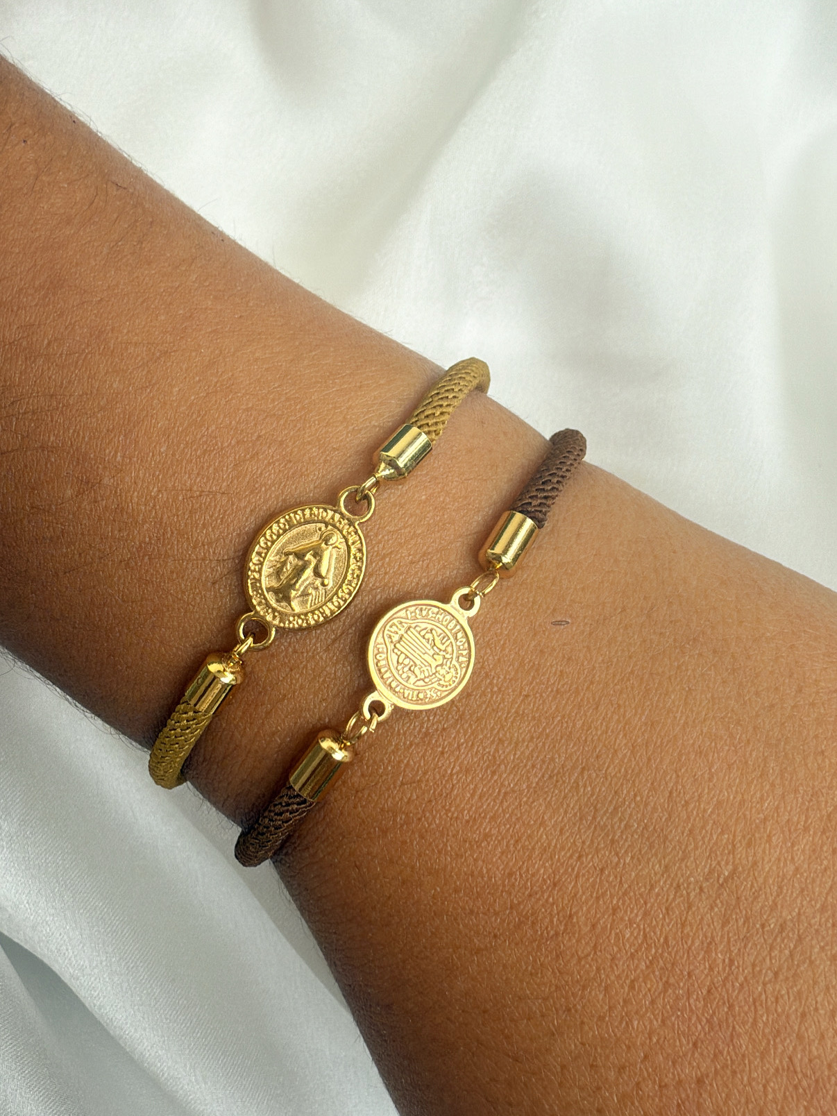 Duas pulseiras de cordão trançado com medalhões dourados em relevo no pulso.