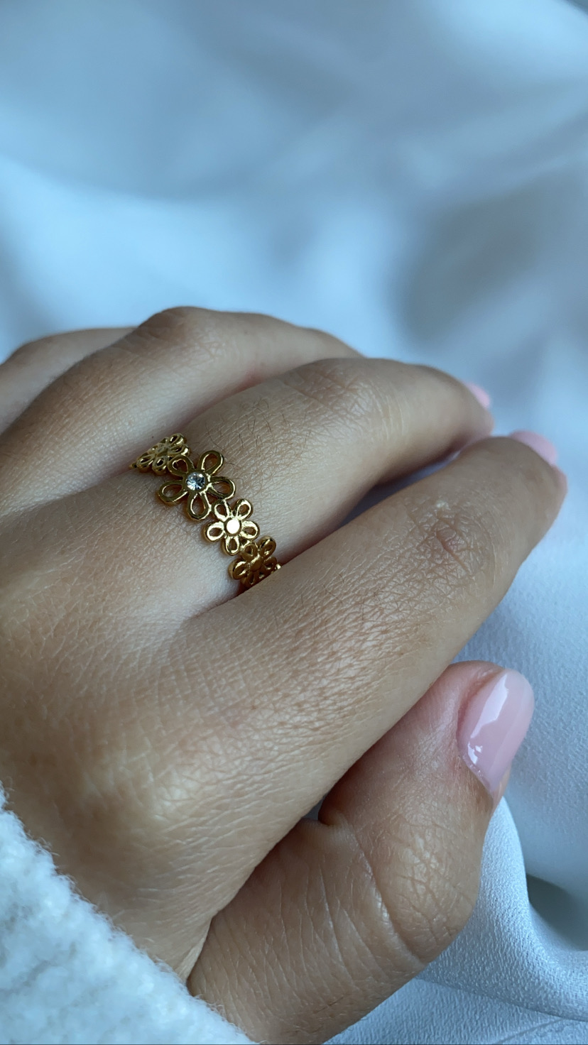 Anel dourado com flores e pedra clara no dedo médio, unhas rosa claro, fundo branco