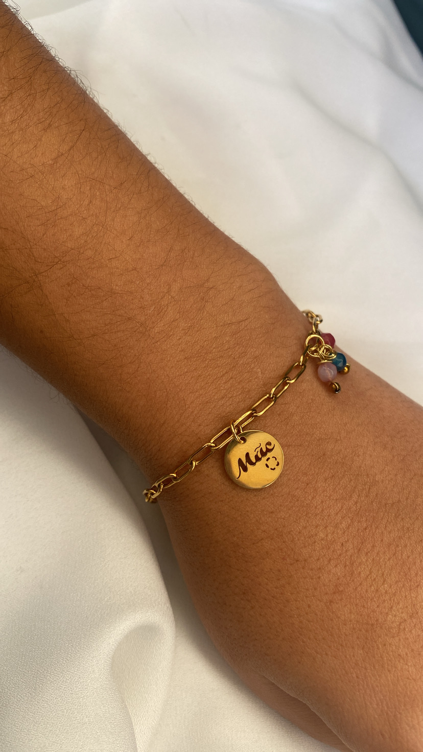 Pulseira dourada com pendente 'Mãe' em pulso sobre tecido branco