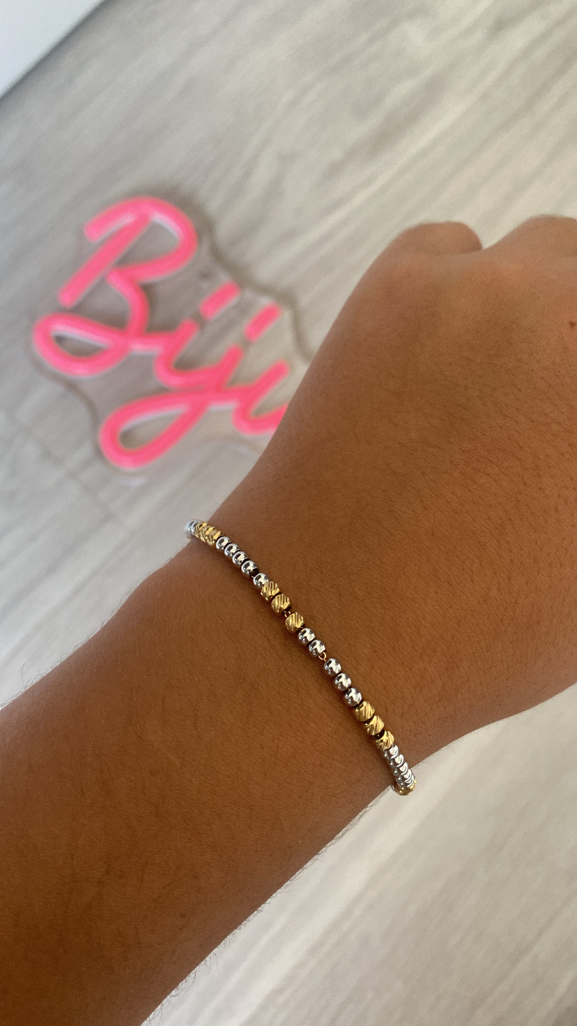 Pulseira fina dourada e prateada num pulso com fundo claro e texto rosa desfocado