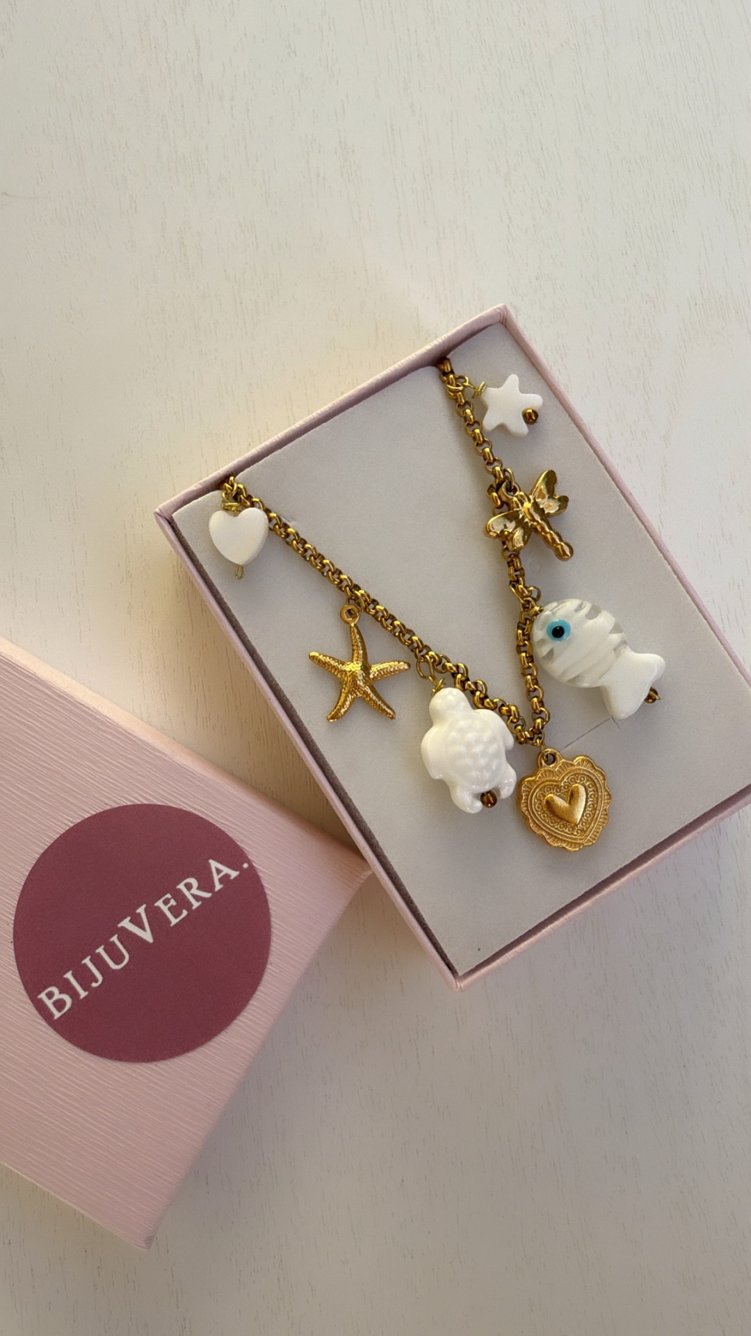 Colar dourado com vários pingentes brancos e dourados dentro de caixa rosa BIJUVERA