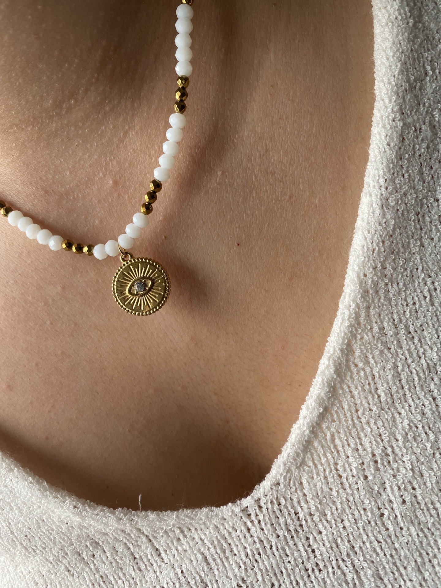 Colar de contas brancas e douradas com pendente de olho dourado sobre pele e camisola bege.