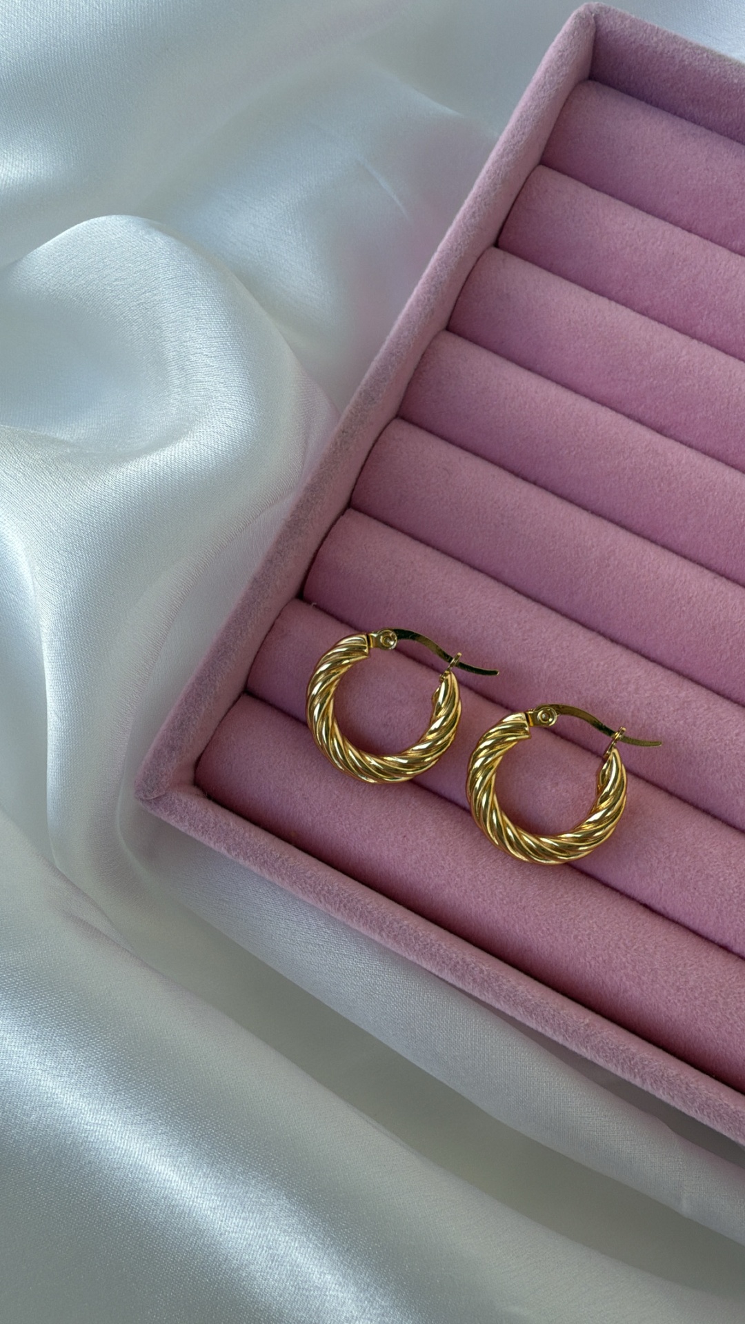 Brincos de argola dourada em caixa rosa sobre tecido branco acetinado
