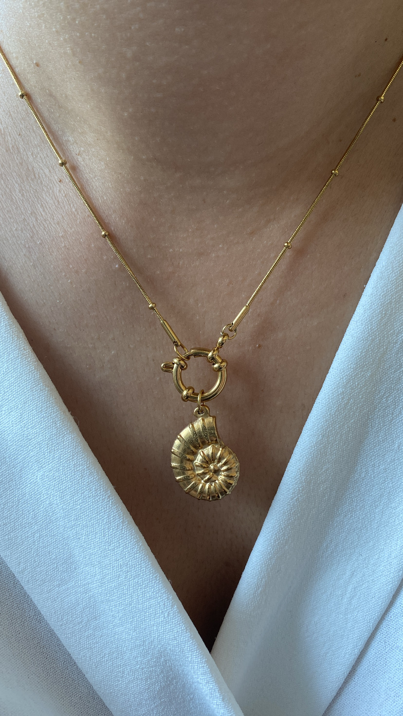Colar dourado com pendente em forma de concha espiral sobre pele e vestido branco