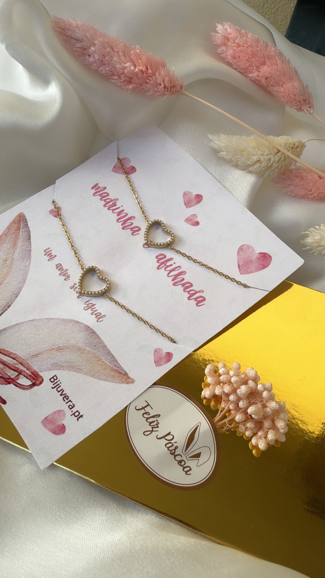 Pulseiras douradas com coração e embalagem com textos em cartão branco sobre tecido acetinado creme e flores secas rosa e creme
