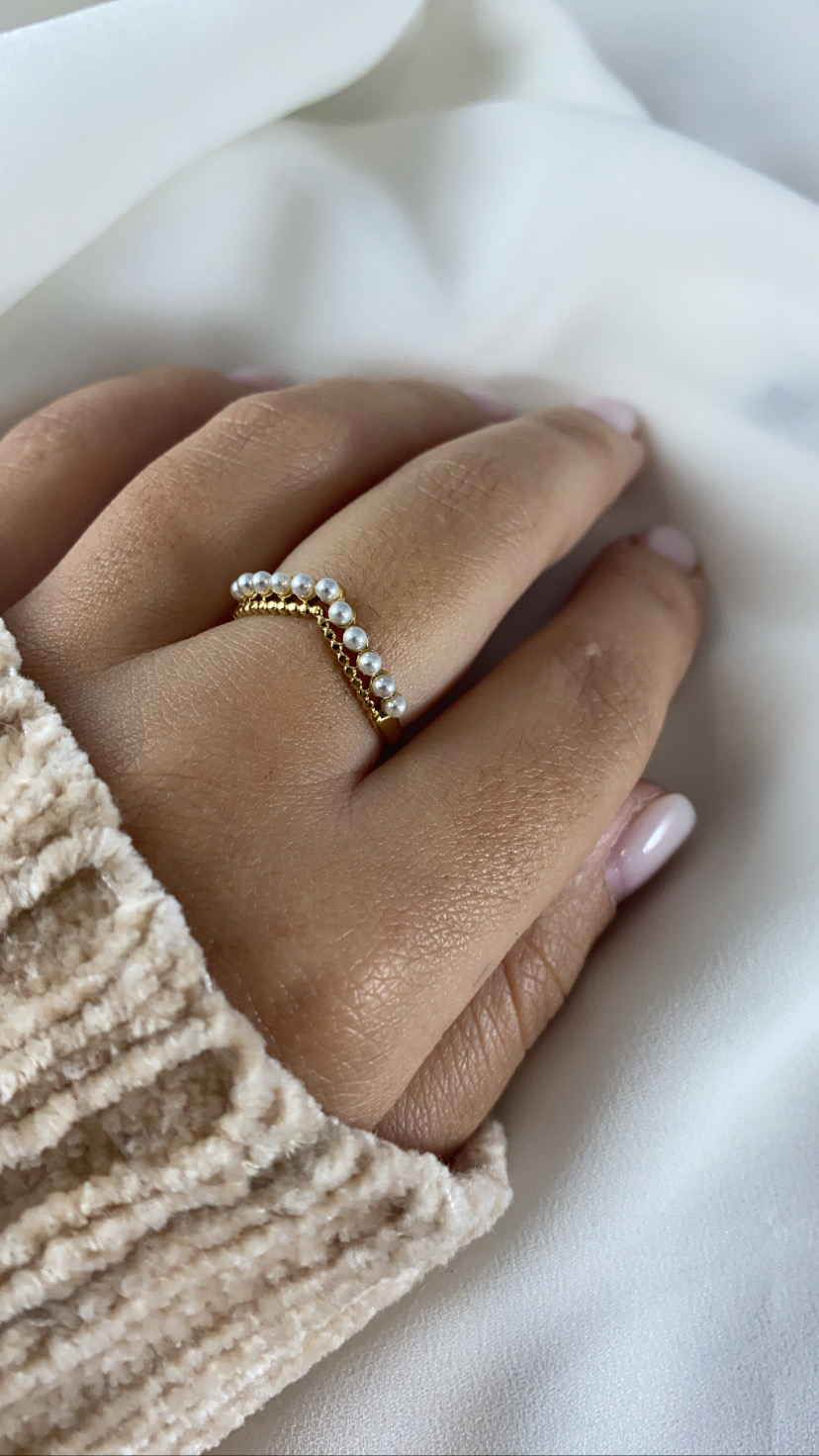 Anel dourado com pérolas pequenas no dedo médio, mão com unhas pintadas, fundo de tecido branco e malha bege.