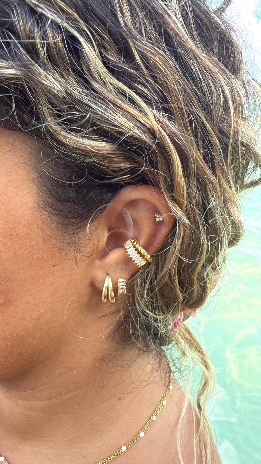 Orelha feminina com brincos dourados variados e cabelo com mechas loiras junto a água