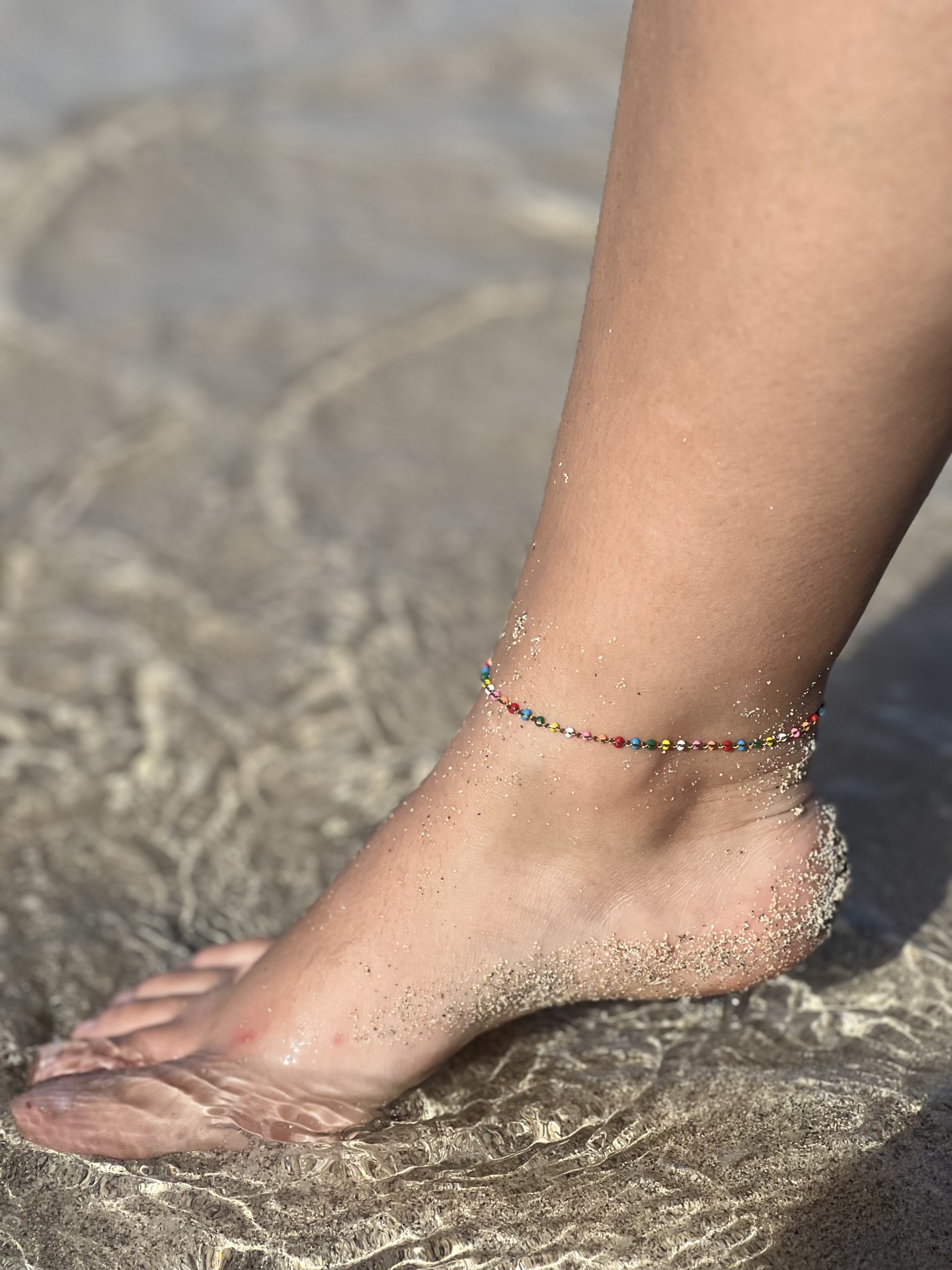 Tornozeleira colorida em pé com areia na praia e água transparente.