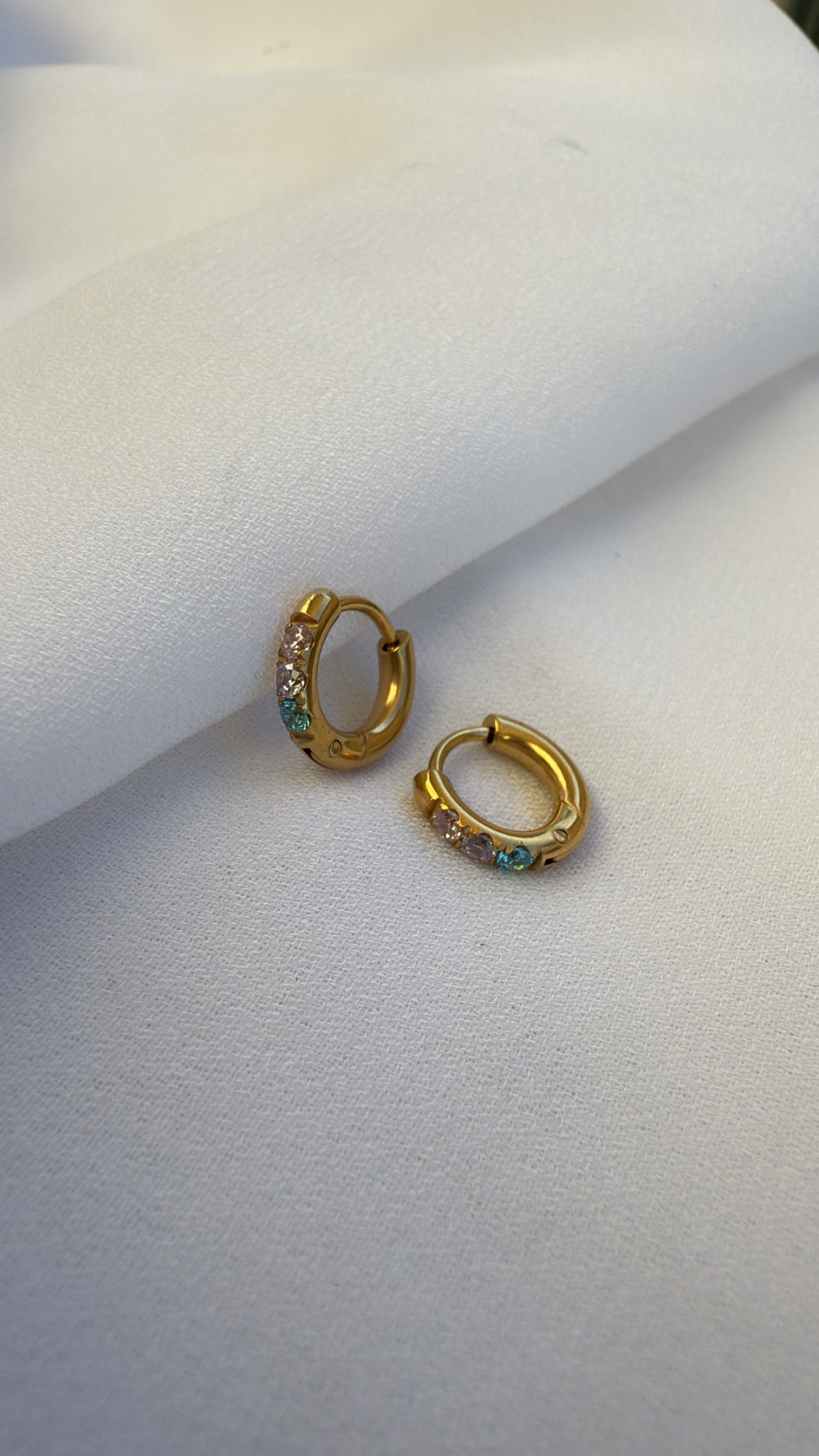 Brincos pequenos em argola dourados com pedras coloridas sobre tecido branco